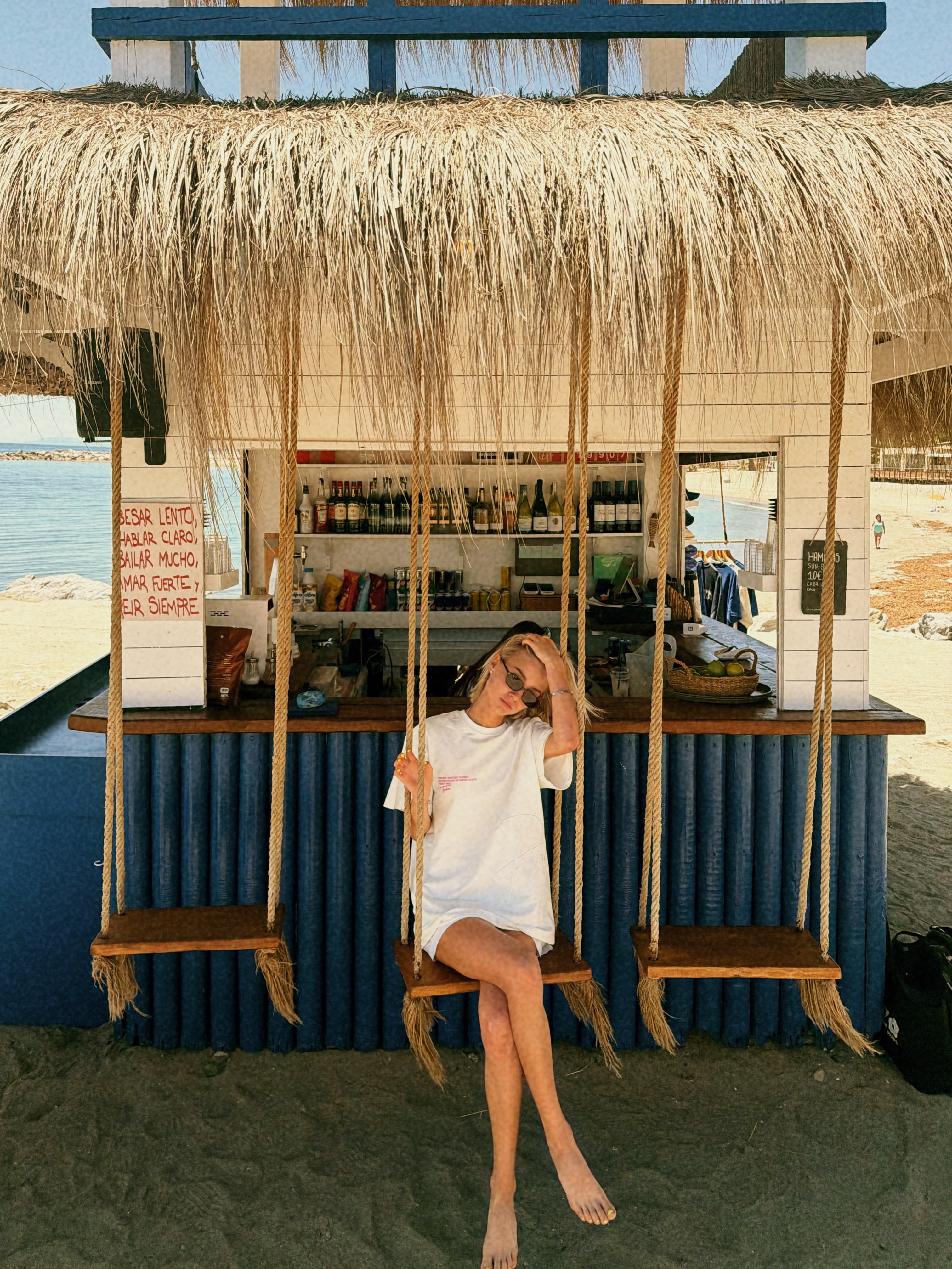 A woman sits barefoot on a wooden swing in front of a beach bar with a thatched roof and blue corrugated walls, wearing a loose white dress.