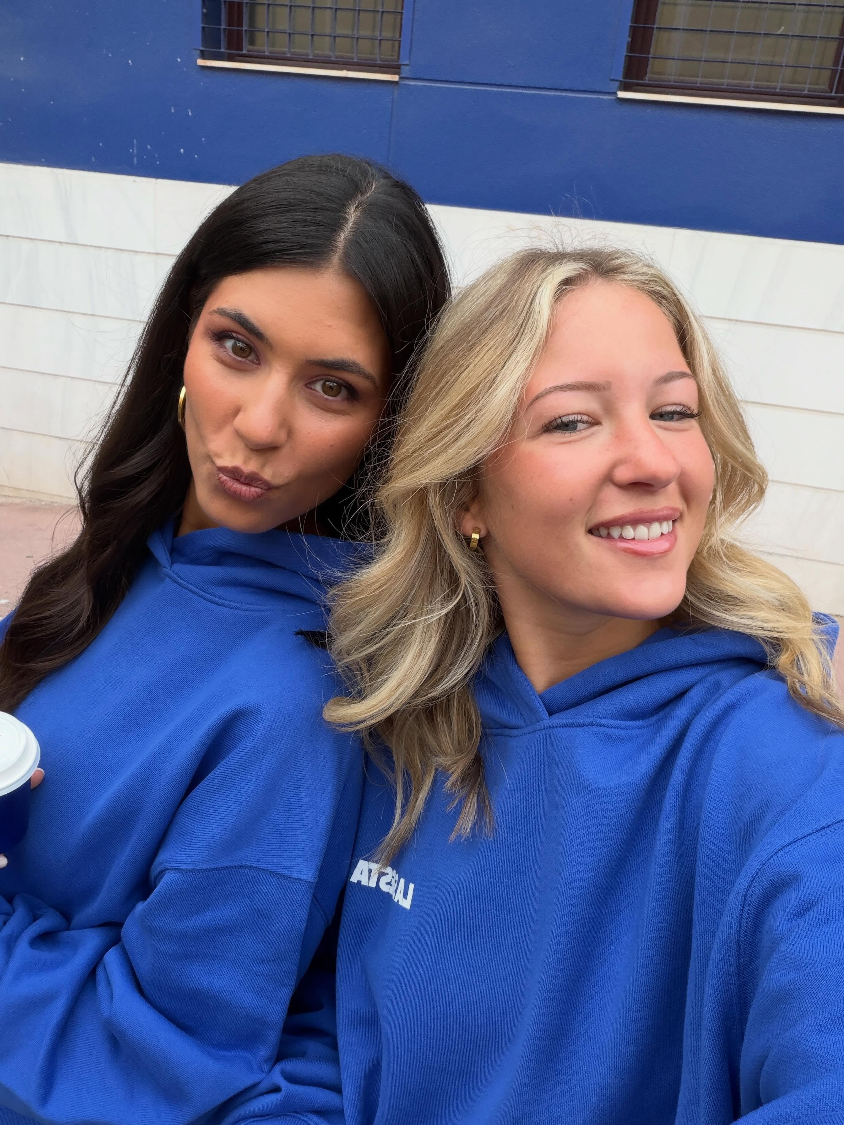 Two women posing together in matching royal blue hoodies with white text on the chest, set against a blue and white building wall.